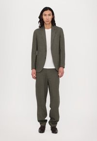 Olive green suit with a textured finish, single-button blazer, loose-fit trousers, and a white t-shirt underneath. Black loafers complete the look.