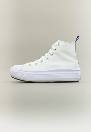 Baskets montantes blanches avec semelle épaisse, lacets blancs, œillets métalliques et petite languette violette au talon sur fond beige.