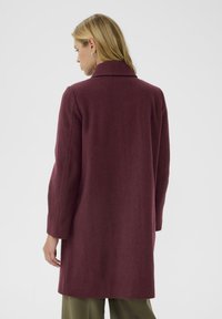 Blonda sieviete, kas stāv pretim, ģērbta garā bordo mētelī un olīvzaļās biksēs uz vienkrāsaina baltā fona.