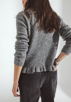 Suéter gris de punto con acabado texturizado, mangas tres cuartos y dobladillo con volantes, combinado con jeans oscuros. Diseño sencillo y funcional.