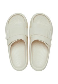 Des sandales en caoutchouc de couleur beige, dotées d'une semelle texturée, d'une large sangle élastique et d'une forme arrondie. Surface lisse avec de petites perforations.