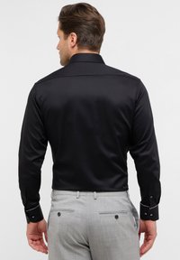 Zwarte satijnen overhemd met knopen en een klassieke kraag, lange mouwen en contrasterende witte accenten op de manchetten. Draagt een grijze broek.