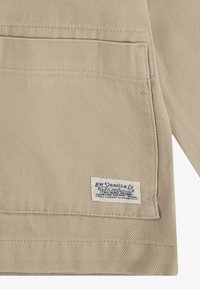 Beiger Stoff mit einer strukturierten Oberfläche, ausgestattet mit einer aufgesetzten Tasche und einem weißen Etikett mit blauer Schrift von Levi Strauss & Co.