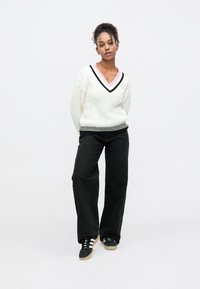 Chunky hvid strikket sweater med V-udskæring, sorte og pink accenter, kombineret med løse sorte bukser og sorte sneakers.