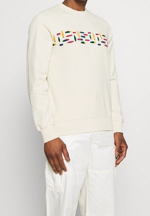 Mann trägt einen cremefarbenen Pullover mit einem mehrfarbigen abstrakten Muster über der Brust und weißen Cargo-Hosen, steht vor einem weißen Hintergrund.