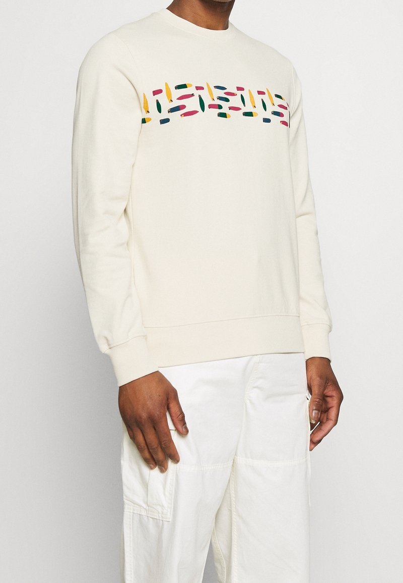 Homme portant un sweat-shirt crème avec un motif abstrait multicolore sur la poitrine et un pantalon cargo blanc, debout devant un fond blanc.