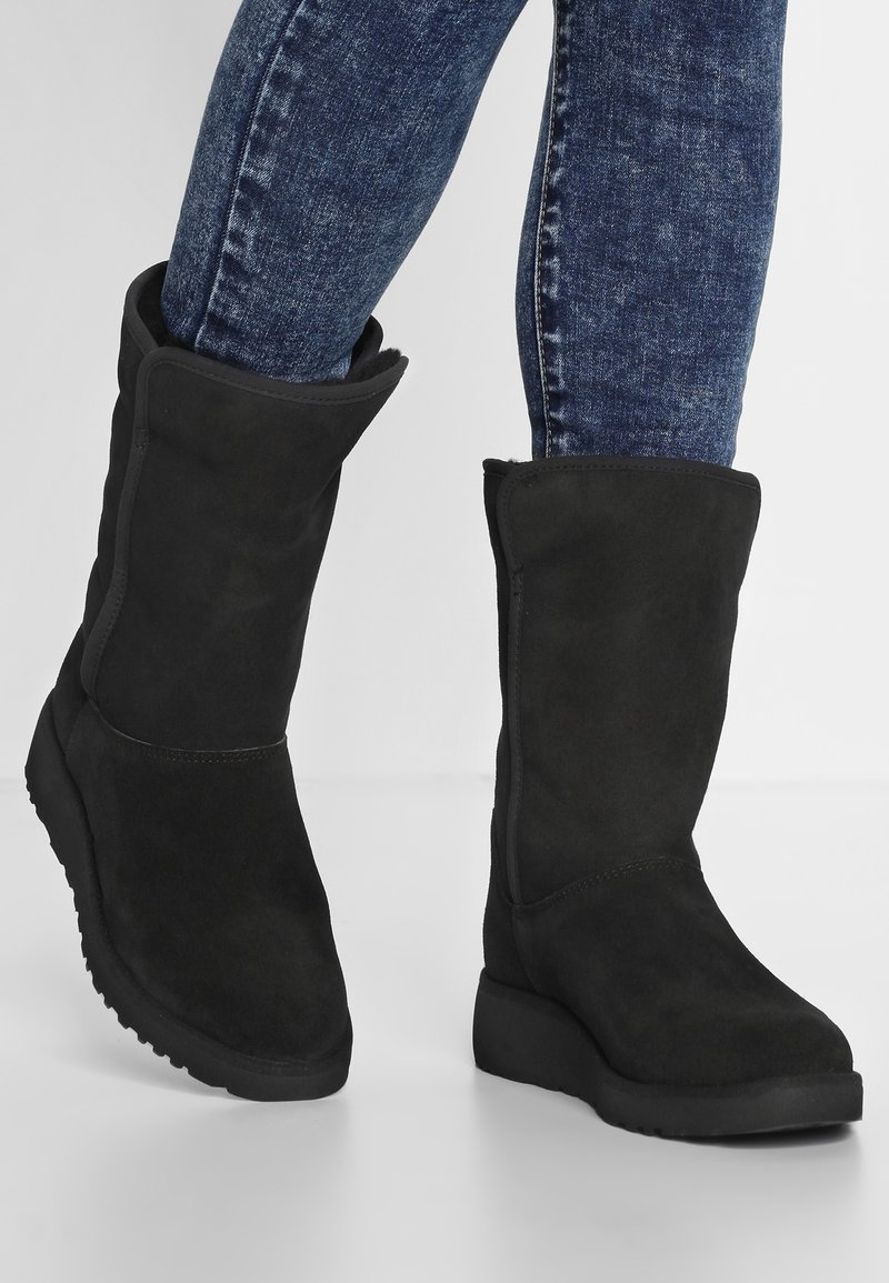 Czarne zamszowe kozaki do połowy łydki z płaską gumową podeszwą, wyposażone w mankiet z podwinięciem i bezszwowy design. Akcenty stanowią przeszycia na krawędziach.