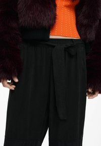 Čierne nohavice s detailom na zaväzovanie v páse, skombinované s textúrovaným bordovým kožušinovým kabátom a oranžovým pleteným topom s malými bielymi bodkami.