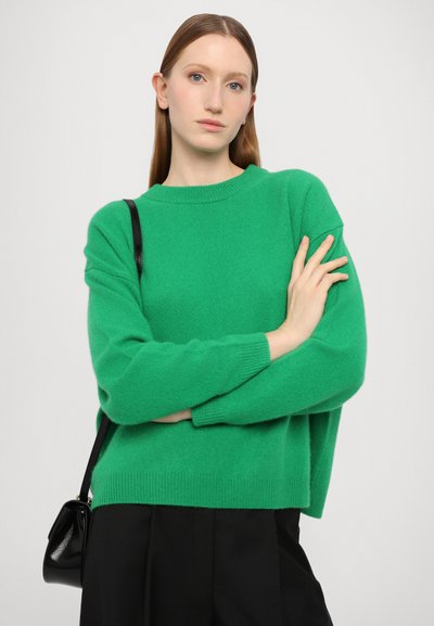 Green knit sweater with a relaxed fit, round neckline, and long sleeves. Paired with black trousers and a small black handbag.