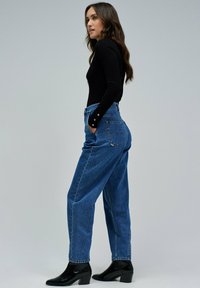 Vaqueros de mezclilla azul de tiro alto con un ajuste relajado, dobladillo enrollado y bolsillos laterales. Combinados con una blusa negra de manga larga ajustada y botas de tacón.