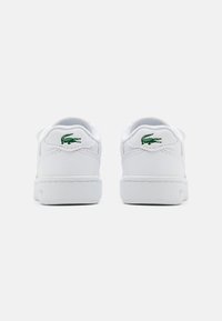 Fehér sportcipő textil és szintetikus felső réteggel, zöld Lacoste logóval a saroknál, lapos fehér gumitalppal és párnázott szegéllyel.