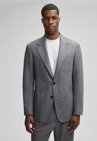 Grauer Wollblazer mit Reverskragen, zwei Fronttaschen und schwarzen Knöpfen, getragen über einem weißen T-Shirt. Texturiert und tailliert geschnitten.