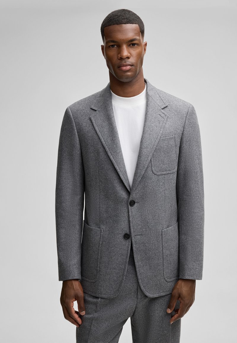 Blazer grigio in lana con colletto a punta, due tasche frontali e bottoni neri, indossato sopra una maglietta bianca. Tessuto testurizzato e vestibilità su misura.