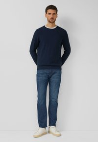 Man staat tegen een muur, gekleed in een marineblauwe trui, blauwe spijkerbroek en witte sneakers, met de handen achter de rug en een neutrale uitdrukking.