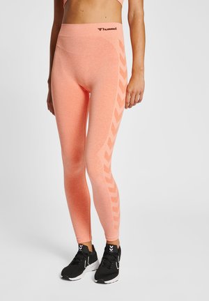 Kvinde iført koralfarvede sømløse leggings med sidet chevronmønster og sorte atletik sko med hvide såler, stående mod en hvid baggrund.