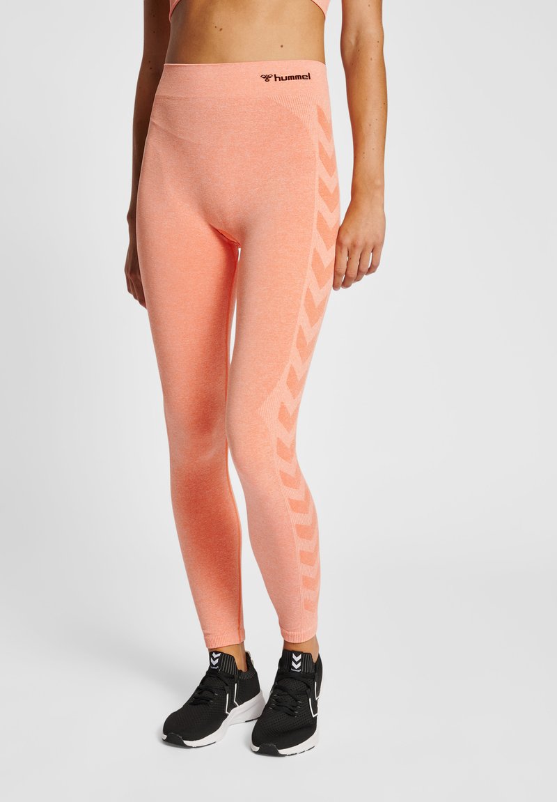 Vrouw draagt koraal kleurige naadloze leggings met een zijwaarts chevronpatroon en zwarte sportieve schoenen met witte zolen, staand tegen een witte achtergrond.