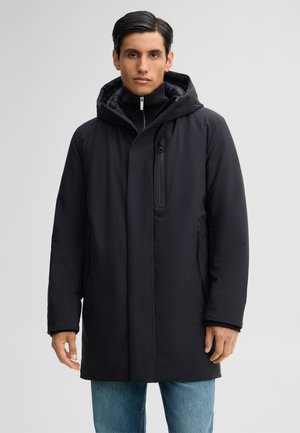 Man wearing a black hooded winter coat with zipper pockets, standing against a plain light background.