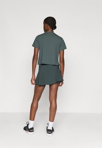 Sötétzöld aktív sportöltözet szett, amely egy rövidített pólóból és egy bővülő szoknyából áll, fekete sportcipőkkel és fehér bokazoknival párosítva.