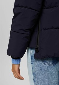 Person iført en sort pufferjakke med en blå sweaterærme synlig og lyseblå denimjeans, hånden afslappet ved siden af.