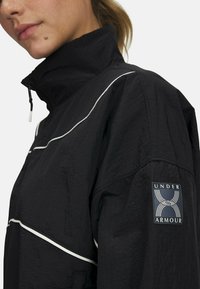 Čierna ľahká bunda s vysokým golierom, ktorá má biele lemovanie pozdĺž ramenných švov a logo Under Armour na rukáve.