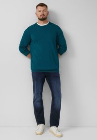 Tiefgrüner Rundhalsausschnitt-Pullover mit langen Ärmeln, kombiniert mit dunklen Jeans und weißen Sneakers. Glatte Textur mit gerippten Bündchen und Saum.