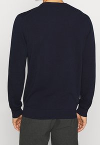 Mann in einem schlichten dunkelblauen Langarmpullover und dunklen grauen Hosen, von der Rückenmitte aufwärts vor einem schlichten Hintergrund gezeigt.