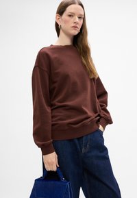 Sudadera marrón de corte holgado con puños acanalados, combinada con pantalones de mezclilla oscuros. Lleva un bolso tote estructurado azul. Diseño minimalista.