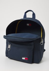 Zaino navy con un compartimento principale e una tasca frontale, con un rivestimento giallo e una patch con il logo "TOMMY JEANS". Fodera con cerniere argento.