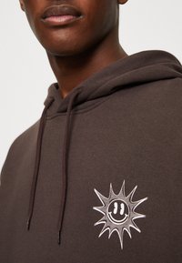 Hoodie marron avec cordon de serrage, présentant un design de rayons de soleil blanc et un visage souriant sur la poitrine gauche. Matière douce avec une coupe décontractée.