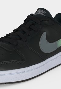 Μαύρο αθλητικό παπούτσι Nike με γκρι λογότυπο swoosh, μαύρα κορδόνια και λευκή σόλα με στίγματα, απεικονίζεται σε κοντινή άποψη από την μπροστινή πλευρά.