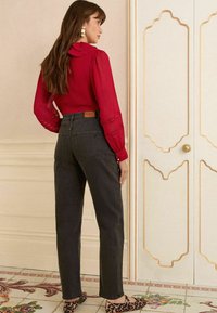 Rdeča bluza z dolgimi rokavi in volančkom, kombinirana z temno jeans hlačami visoko pasu in balerinkami z leopardjim vzorcem, na nevtralni podlagi.