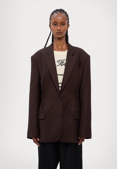 Pruuni mustriga ülerõivas, mis on suurem, struktureeritud õlgadega, ühe nupuga kinnitusega ja kahe esikotiga, kihtide kaupa kantud kreemja sviidri peal.