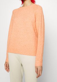 Jersey de color naranja claro de punto con cuello redondo y puños acanalados, combinado con pantalones beige texturizados. Textura suave y corte relajado.