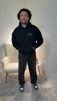 Mann mit lockigem Haar, der einen schwarzen Hoodie, dunkle Jeans und Turnschuhe trägt, steht drinnen neben einem weißen Sessel und einem kleinen Tisch mit Büchern.