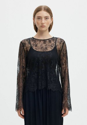 Une femme porte une robe noire avec un haut en dentelle florale transparente et des manches longues, sur un fond clair uni.