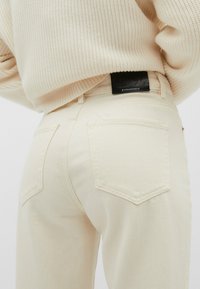 Jean crème avec une texture lisse, dotés de deux poches arrière et d'une étiquette de marque noire à la taille, associés à un pull léger en tricot.