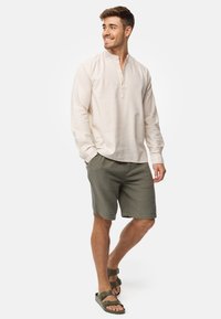 Camicia beige a maniche lunghe con dettaglio a bottoni, abbinata a pantaloni corti verdi con texture. Il modello indossa sandali marroni, in piedi su uno sfondo neutro.
