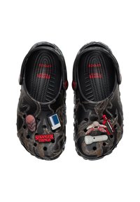 Een paar zwarte Crocs met grijze en bruine strepen, versierd met Stranger Things-thema charmes, waaronder een cassettebandje en een Demogorgon-figuur.