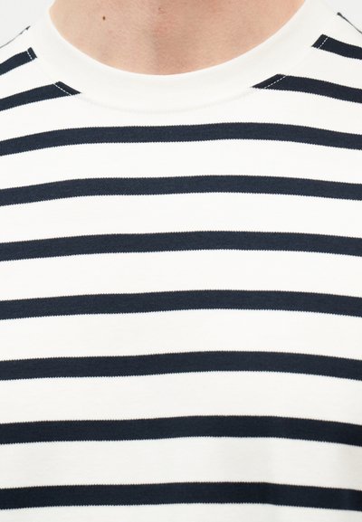 Κοντινή λήψη ενός ατόμου που φοράει λευκό πουκάμισο με ομοιόμορφες οριζόντιες μαύρες ρίγες και στρογγυλεμένο λαιμόκοψη.