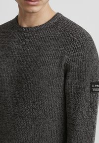 Dark gray knitted sweater with a textured pattern and a crew neckline. Features a black label on the left sleeve with white text.