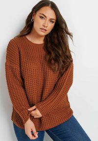 Sweter w kolorze rdzy z dzianiny o teksturowanym, waflowym wzorze i luźnym kroju. Posiada długie rękawy i okrągły dekolt, zestawiony z niebieskimi dżinsami.