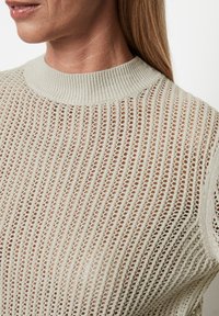 Kobieta ubrana w beżowy sweter z otwartym splotem, z prążkowanym okrągłym dekoltem i widoczną skórą pod spodem.