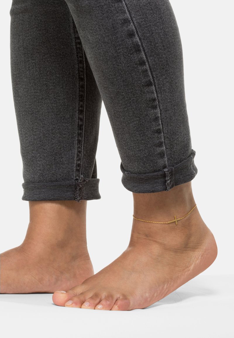 Cavigliera d'oro con charm a croce indossata sopra un piede nudo, abbinata a jeans grigi scuri arrotolati alla caviglia. Texture liscia e design casual.