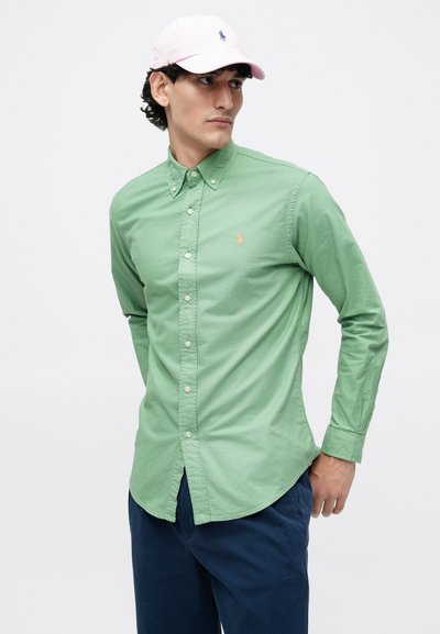 Homem com uma camisa verde abotoada, calças azul-marinho e um boné rosa claro olhando para o lado contra um fundo branco simples.