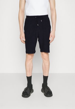 Pantaloni corti in misto cotone di colore blu navy con vita elastica e cordoncino, caratterizzati da una vestibilità comoda e una texture liscia. Indossati con calze nere e sandali.