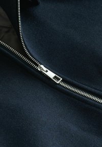 Marineblaue Stoff mit glatter Textur, ausgestattet mit einem silbernen Reißverschluss und einem Stehkragen-Design. Der Reißverschluss ist auffällig präsentiert.