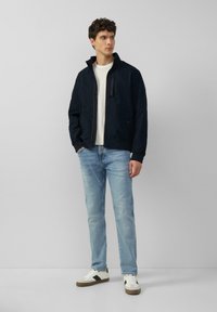 Marine bomberjack met staande kraag, ritssluitingen aan de voorkant en zwarte accenten, gecombineerd met lichtblauwe jeans en witte sneakers met bruine zolen.