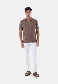 Polo shirt marrone a manica corta con colletto, chiusura a bottoni, abbinato a pantaloni bianchi e sandali marroni su uno sfondo semplice.