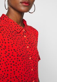Červená košile s krátkým rukávem a černou tečkovanou vzorem, s knoflíkovým zapínáním a límcem. Vyrobena z texturované tkaniny. Doplněna oranžovými knoflíky.