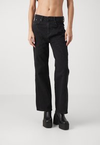 Mujer vestida con pantalones negros de talle alto con paneles cosidos y botas de plataforma negras, de pie contra un fondo claro y liso.